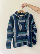 Buzo estilo mexicano con capucha, de tejido grueso a rayas horizontales en tonos azul, celeste, gris y negro. Tiene un bolsillo tipo canguro en el frente y cordones ajustables en el cuello.