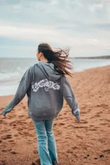 Hoodie gris con capucha y cordones ajustables, de corte holgado y manga larga. Presenta un bordado de flores blancas en la espalda y un pequeño bordado en la manga.