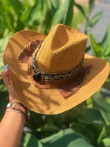 Sombrero de ala ancha tejido en hilo de coco y sintético, color marrón claro. La copa está cubierta con encaje naranja quemado con patrón floral. Tiene una cinta de cuero marrón oscuro y un aplique decorativo de hoja de gamuza con incrustaciones de pedrería.