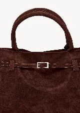 Cartera marrón de gamuza con doble asa corta, correa con hebilla decorativa y cierre superior.