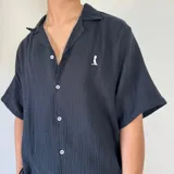 Camisa negra de muselina con manga corta, corte oversize, cuello cubano, cierre con botones blancos y logo bordado en blanco en el pecho.