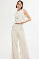 Pantalón de corte holgado color beige, en tejido texturizado con pliegues adelante y atrás. Modelo de tiro alto con elástico oculto en la cintura y piernas anchas. Bolsillos delanteros decorativos con botón de metal con diseño.