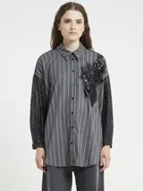Camisa de manga larga con rayas verticales grises y blancas, cuello camisero y cierre frontal de botones oscuros. Las mangas son de encaje negro, y el frente presenta una aplicación decorativa de lentejuelas y cuentas negras en forma de flor o estrella.