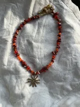 Collar de piedras de color naranja y dije de sol plateado. Las piedras son de diferentes tamaños y formas. El dije de sol es de bronce y tiene un diámetro de 2.5 cm.