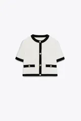 Blazer blanco de tejido de punto con cuello redondo, manga corta, bolsillos laterales y cierre frontal con botones metálicos dorados. Presenta ribetes negros en cuello, mangas, cierre y bolsillos.