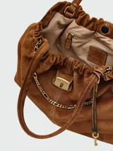 Bolso tipo hobo o bucket de gamuza marrón, con frunce en la parte superior y detalles de costura en contraste. Cuenta con una cadena dorada decorativa y un dije metálico con logo.