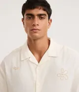 Camisa de hombre con corte boxy, cuello inglés y abotonado delantero. Es de color crema y presenta bordados sutiles de motivos veraniegos en tono naranja claro.