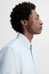 Camisa celeste a rayas verticales, de corte regular fit, confeccionada en tejido de algodón. Tiene cuello abotonado, manga larga con puños abotonados, bolsillo de parche en el pecho y cierre frontal con botones.