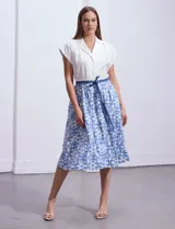 Vestido midi camisero de algodón con estampado floral en tonos azules y blancos. Tiene cuello solapado, cierre frontal con botones, manga corta, cintura elastizada con lazo de ajuste y bolsillos laterales.