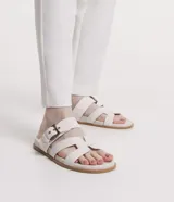 Sandalias planas de cuero sintético color blanco, con puntera redonda y dos tiras, una con hebilla metálica y otra en forma de H.