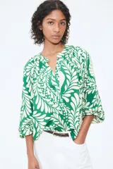 Blusa de tejido liviano color verde con estampado de hojas blancas, cuello corto fruncido y tapeta de botones. Mangas globo largas con volumen de corte raglán y puños finos abotonados.
