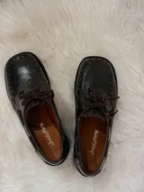 Zapato tipo mocasín de cuero color chocolate, con suela plana de goma negra, cordones al tono y costuras decorativas visibles en el borde superior.