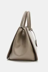 Bolso de mano tipo satchel o tote pequeño, color topo o gris pardo, con doble asa superior y cierre principal por cremallera. Presenta un colgante rectangular de cuero sintético colgando de una de las asas.