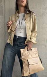 Sobrecamisa beige de gamuza con cuello camisero, cierre frontal con botones y mangas largas con puños abotonados. Presenta bolsillos de parche en el pecho y detalle de calado en el ruedo.