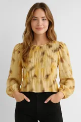 Blusa color amarillo con estampado abstracto en tonos marrones, de poliéster con frunces, lurex, punto smock y volados. Tiene mangas largas abullonadas con elástico en los puños y cuello redondo con elástico.