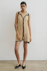 Vestido corto color beige con cuello camisero, sin mangas, con botones frontales y ribete negro en cuello, sisas y ruedo.