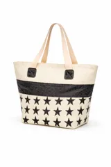 Bolso tipo tote bag de tela color beige con estampado de estrellas negras en la parte inferior y una franja horizontal negra brillante en el medio. Tiene asas de tela color beige claro y detalles de cuero sintético negro en los puntos de unión de las asas.