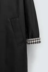Gabardina negra de corte recto con cuello solapa, cierre frontal con botones, bolsillos laterales y mangas largas. Interior con forro a cuadros. Edición limitada.