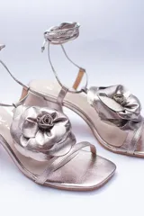 Sandalias de cuero color champagne, con taco cuadrado, tira delgada sobre los dedos y tobilleras para atar. Presentan un aplique de flor de cuero en la capellada.