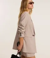 Blazer largo color beige, de corte recto, con cuello solapa, cierre frontal con doble botón, bolsillos tipo ojal y mangas largas con botones decorativos en los puños.