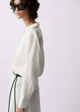 Camisa blanca de lino con cierre de botones en la parte delantera, mangas largas con puños abotonados, cuello camisero y corte recto. Incluye cinturón trenzado negro.