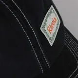 Gorra de béisbol negra con costuras de contraste en hilo blanco. Presenta un parche rectangular frontal con el texto "Rivvia" en naranja y un borde decorativo de flores blancas.
