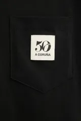 Camiseta negra de algodón con cuello redondo y manga corta. Tiene un bolsillo de parche en el pecho con el logo del 50 aniversario.