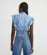 Camisa cropped de jeans celeste, sin mangas, con cuello alto, cierre frontal con botones, volantes en cuello y laterales, y elástico en la cintura.