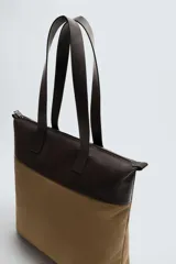 Bolso tipo shopper de tela color marrón con detalles en cuero color marrón oscuro. Tiene doble asa de mano de cuero y cierre principal con cremallera.