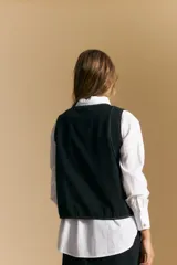 Chaleco negro de vestir con cuello solapa y cierre frontal de botones. Presenta costuras contrastantes blancas que definen el diseño y bolsillos delanteros. Se combina con una camisa blanca de manga larga.