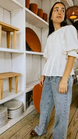 Blusa de color natural confeccionada en tejido 100% algodón. Presenta un cuerpo delantero fruncido tipo smock, vuelo peplum en la cintura y mangas globo 3/4 con volados en los puños.