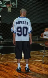 Camiseta estilo jersey de fútbol americano, color blanco, con cuello en V ribeteado en azul. Presenta el número "90" grande en el frente en azul marino con borde blanco. Tiene bordados en el pecho como "Lx." y "Ravers", y números pequeños en las mangas. Los puños tienen franjas azules y negras.