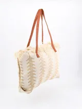 Bolso tipo tote color beige con diseño geométrico en blanco y flecos en la parte superior. Tiene doble asa de hombro marrón.