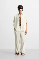 Pantalón relaxed fit de color beige con finas rayas verticales, confeccionado en una mezcla de algodón y cáñamo. Presenta cintura elástica, bolsillos laterales, un bolsillo trasero de plastrón y cierre frontal con botones.