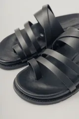 Sandalia plana negra de cuero con diseño de tiras anchas entrecruzadas sobre el empeine y una tira que separa el dedo gordo. Suela plana y gruesa.