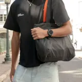 Bolso tote negro de tela resistente al agua, con asas de correa negras con borde naranja. Tiene cierre principal, bolsillos laterales (uno con cierre), seis bolsillos internos, diseño plegable y capacidad de 14,5 litros.