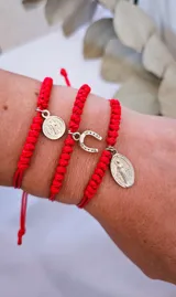 Set de tres pulseras rojas de hilo trenzado con dijes de metal plateado: medalla de San Benito, herradura y medalla de la Virgen María.