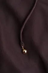 Vestido corto color marrón oscuro, estilo camisero, con cuello mao con ribete de volado y tapeta de botones con el mismo detalle. Posee mangas largas con puños sencillos de volado y un cinturón fino extraíble para ajustar la cintura. La falda tiene un fruncido escalonado que le da vuelo.