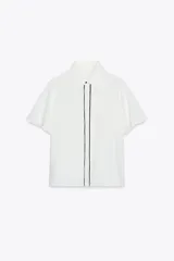 Camisa blanca de corte recto, con cuello solapa y mangas cortas. Presenta vivos negros a contraste en el frente y aberturas laterales en el ruedo. Cierre frontal con botones ocultos.