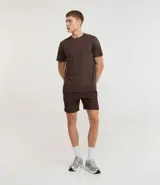 Bermuda deportiva masculina color marrón oscuro, confeccionada en poliéster reciclado certificado RCS. Presenta cintura elástica con lazo y un estampado abstracto en tonos claros en la pierna izquierda.
