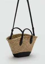 Bolso tipo canasta de paja trenzada color beige con base y asas cortas de cuero sintético negro.