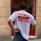 Camiseta blanca de algodón con estampado en la espalda de una lancha con una mujer y el texto "Locally Hated" en rojo.