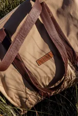 Bolso tote color beige con asas de cuero marrón, cierre superior y bolsillo interno.