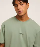 Remera verde salvia de corte relaxed, confeccionada en algodón más sostenible. Presenta cuello redondo, mangas cortas y estampa frontal con lettering y árboles.