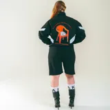 Campera deportiva negra de nylon liviano con paneles diagonales blancos en contraste. Presenta un estampado frontal discreto y un gran gráfico en la espalda que incluye una silla naranja con un gato blanco durmiendo y la frase "Sin siesta no hay fiesta".