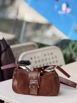 Cartera marrón con correa al hombro, hebilla decorativa y detalles de tachas.