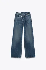 Pantalón de jean celeste de tiro alto, corte wide leg y largo hasta el tobillo. Presenta un diseño asimétrico en la cintura con cierre frontal cruzado con cremallera y botones metálicos.