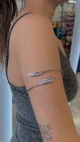 Brazalete plateado para el brazo, con diseño abierto y dos dijes en forma de pluma en los extremos.