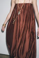Vestido largo marrón de tejido satinado, con escote recto, tirantes finos extraíbles, bolsillos laterales ocultos y bajo asimétrico acabado en globo.