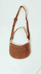 Bolso de hombro tipo media luna de gamuza color marrón, con bordes cosidos a mano en contraste claro. Cuenta con correa ajustable de gamuza y una correa adicional trenzada. El logo de la marca está grabado en la parte inferior central.